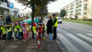 Dzielnicowy z grupą przedszkolaków na chodniku, tłumaczy zasady przechodzenia przez jezdnię na przejściu dla pieszych.