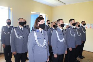 15 policjantów ubranych w mundury galowe ustawieni w trzech szeregach oczekują na wręczenie awansów na wyższe stopnie policyjne.