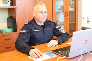 I Zastępca Komendanta Powiatowego Policji w Łasku młodszy inspektor Robert Krawczyk w gabinecie, w umundurowaniu służbowym siedzi przy stoliku przed monitorem laptopa prowadząc zajęcia online z młodzieżą.