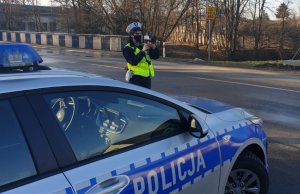 Na poboczu przy radiowozie oznakowanym stoi umundurowana policjantka ruchu drogowego zwrócona w kierunku jezdni i dokonuje pomiaru prędkości.