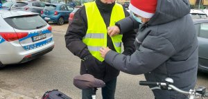 Pracownik Wojewódzkiego Ośrodka Ruchu Drogowego zapina kamizelkę odblaskową mężczyźnie. W tle oznakowany radiowóz policyjny.