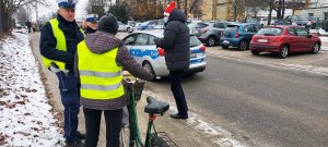 Policjantka z policjantem Wydziału Ruchu Drogowego oraz pracownik Word w czapce Mikołaja rozmawiają z rowerzystką która ma na sobie założoną kamizelkę odblaskową.