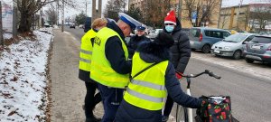 Policjantka z policjantem Wydziału Ruchu Drogowego oraz pracownik Word w czapce Mikołaja rozmawiają z dwiema rowerzystkami, które mają na sobie założone rozdane kamizelki odblaskowe.