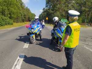 Policjantka stoi twarzą do kierujących policyjnymi motocyklami. Policyjne motocykle mają włączone sygnały świetlne koloru niebiesko czerwonego.