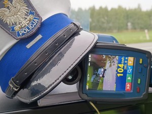 Czapka policjanta ruchu drogowego oraz ręczny miernik prędkości. Na urządzeniu pomiarowym wynik pomiaru - 104 kilometry na godzinę.