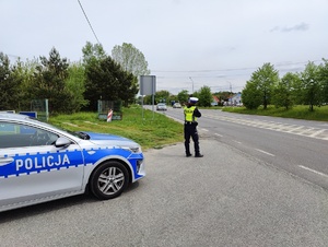 Policjantka stojąca przy policyjnym radiowozie mierzy prędkość pojazdów.