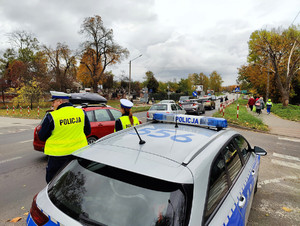 Policjantka z policjantem stoją przy radiowozie i obserwują ruch pojazdów przy cmentarzu w Łasku.