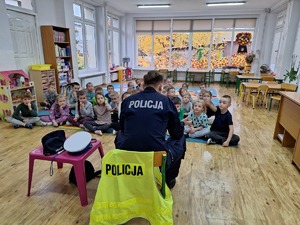 Policjant rozmawia z przedszkolakami o bezpieczeństwie. Dzieci siedzą przed policjantem na dywanie.