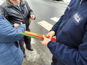 Policjant trzyma w ręce opaski odblaskowe i rozdaje je pieszym.