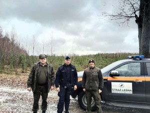 Policjant wraz z dwoma strażnikami leśnymi pozuje do zdjęcia na tle lasu.