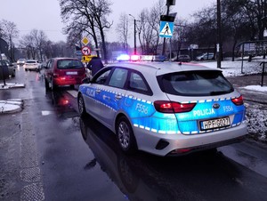 Policyjny radiowóz stoi przed przejściem dla pieszych na włączonych sygnałach świetlnych.