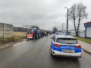 Kolumna ciągników rolniczych ustawiona wzdłuż ulicy kolejowej w Łasku. Przed kolumną na drodze stoi policyjny radiowóz.