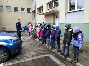 Dzieci stoją przed policyjnym radiowozem. Policjant, który stoi przed nimi opowiada o policyjnym wozie.