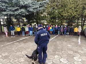 Dzieci stoją przed policyjnym przewodnikiem psa, który prezentuje pokaz tresury.