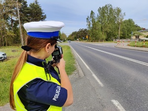Policjantka stoi przy drodze i sprawdza prędkość jadących nią pojazdów.