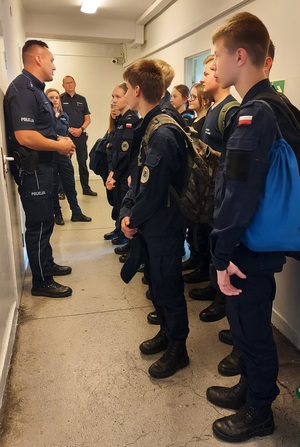Policjant stojący na korytarzu z lewej strony. Z prawej uczniowie z klasy mundurowej.