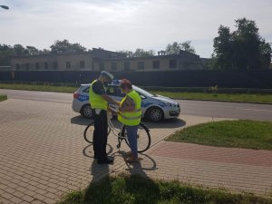 Policjant ruchu drogowego stoi obok rowerzystki na ścieżce rowerowej i pomaga jej zapiąć kamizelkę odblaskową, która została wręczona w ramach akcji Road Safety Days. W tle stoi policyjny radiowóz marki Kia Ceed.