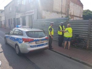 Dwaj policjanci ruchu drogowego stoją na chodniku z rowerzystką. Jeden z nich wręcza kobiecie kamizelkę odblaskową, którą pomaga założyć. Na pierwszym planie stoi ustawiony wzdłuż chodnika policyjny radiowóz kia.
