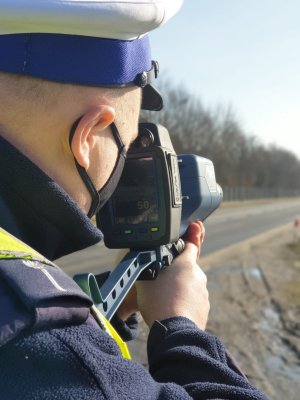 W przybliżeniu umundurowany policjant Wydziału Ruchu Drogowego dokonuje pomiaru z urządzenia TruCAM - przybliżenie na wyświetlacz urządzenia na którym widać pomiar prędkości.