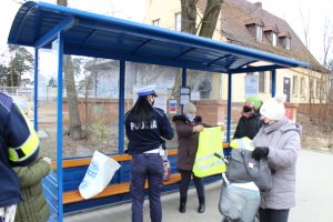 W wiacie przystankowej tyłem stoi umundurowana policjantka, która wręcza kamizelki odblaskowe, obok stoją trzy kobiety, jedna z nich trzyma przed sobą rozłożoną kamizelkę.