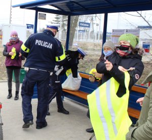 Policjanci Wydziału Ruchu Drogowego na przystanku autobusowym wręczają paniom kamizelki odblaskowe i lizaki, na zdjęciu jest dwójka policjantów tyłem - kobieta i mężczyzna oraz trzy obdarowane panie, jedna z nich na pierwszym planie przymierza kamizelkę.