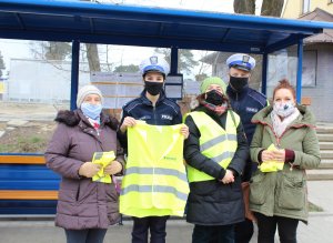 Na tle przystanku autobusowego stoją trzy kobiety, które otrzymały kamizelki, dwie z kobiet trzyma je złożone w rękach, jedna kobieta w środku ma założoną kamizelkę odblaskową. Pomiędzy paniami stoi policjant oraz policjantka z Wydziału Ruchu Drogowego.