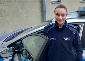 Zdjęcie przedstawia policjantkę w umundurowaniu służbowym stojącą przy radiowozie oznakowanym.