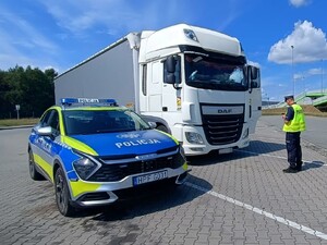 Policjant wydziału ruchu drogowego kontroluje pojazd ciężarowy. Obok pojazdu ciężarowego stoi radiowóz policyjny.