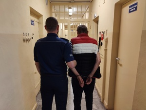 Policjant trzyma za rękę poszukiwanego mężczyznę. Poszukiwany ma założone kajdanki na ręce trzymane z tyłu.