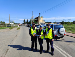 Policjantka Wydziału Ruchu Drogowego pozuje do zdjęcia wraz z dwoma funkcjonariuszami Straży Ochrony Kolei. W tle ustawione pojazdy przed zamkniętym przejazdem kolejowym.