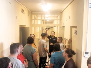 Policjant prezentuje zaproszonym gością wykrywacz metali. Wycieczka znajduje się w pomieszczeniu dla osób zatrzymanych.