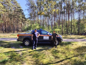 Policjant wraz z funkcjonariuszem Straży Leśnej stoi przy zaparkowanym w lesie radiowozie straży leśnej.
