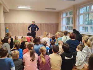 Policjant stoi przed uczniami i rozmawia z nimi o bezpieczeństwie w ruchu drogowym. Uczniowe siedzą przed policjantem na ławkach.