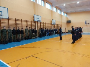 Policjanci oraz uczniowie klas mundurowych stoją w rzędach. Policjantka omawia kolejne ćwiczenia.