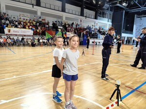 Chłopiec i dziewczynka stoją na lini startu, w tle policjantka - sędzia główny zawodów.