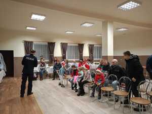 Policjant prowadzi spotkanie poriflaktyczne, opowiada o bezpieczeństwie na drodze. Przed policjantem siedzą dorośli oraz dzieci.