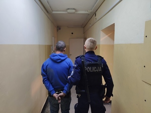 Policjant trzyma zatrzymanego mężczyznę. Mężczyzna ma kajdanki założone z tyłu.