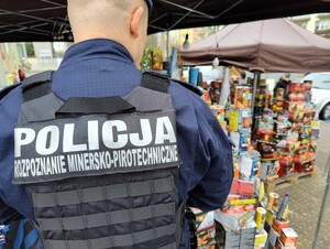 Na kamizelce taktycznej policjanta widać napis Policja rozpoznanie minersko pirotechniczne. W tle widać fajerwerki.