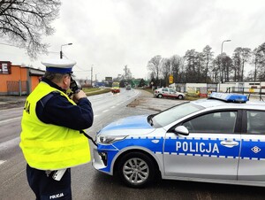 Policjant stoi przed policyjnym radiowozem i kontroluje prędkość pojazdów.
