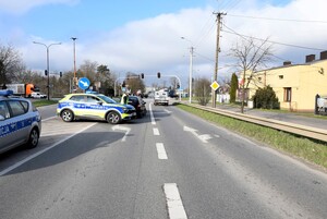Policjant dokonuje oględzin pojazdu sprawcy zdarzejnia. Na drodze stoją dwa radiowozy policyjne oraz auto sprawcy kolizji. W tle odbywa się ruch pojazdów.