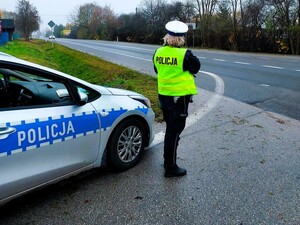 Policjantka kontroluje prędkość pojazdów. Stoi przy zaparkowanym radiowozie policyjnym.
