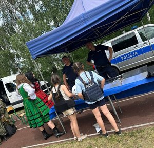 Funkcjonariusze Policji przy stoisku promocyjno - prewencyjny. Za stoskiem rodiowóz policyjny typu bus. Przed stoiskiem osoby.