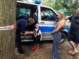 Chłopiec przebrany za policjanta uśmiecha się do zdjęcia. Obok stoi policjantka, która rozmawia z chłopcem.