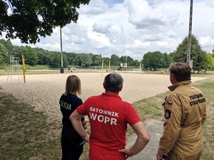 Policjantka wraz z ratowinikiem oraz strażakiem kontrolują kąpielisko miejskie. W tle widać plaże oraz kąpiące się osoby.