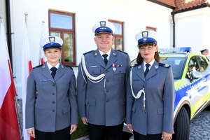 Policjant z policjantkami pozuje do zdjęcia. W tle widać policyjny radiowóz.