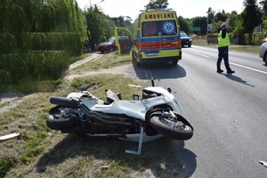 motocykl leżący na poboczu drogi, na środku jezdni funkcjonariusz policji kierujący ruchem, obok motocykla karetka pogotowia.