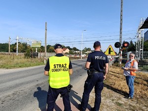 Polcjant wraz z funkcjonariuszem Straży Ochrony Kolei stoi przed zamkniętym przejazdem kolejowym.