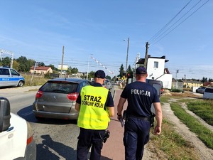 Polcjant wraz z funkcjonariuszem Straży Ochrony Kolei idą chodnikiem w kierunku otwierającego się przejazdu kolejowego.