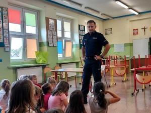 Policjant stoi przed siedzącymi dziećmi i mówi im o zasadach bezpieczeństwa.