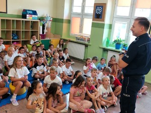 Policjant stoi przed siedzącymi dziećmi i mówi im o zasadach bezpieczeństwa.